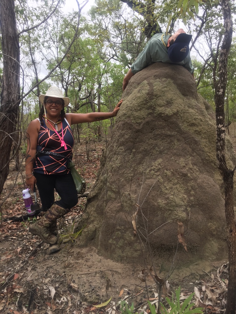 Jessica Ware stands next to a giant termite mound that is taller than her while her colleague lays on top of it.