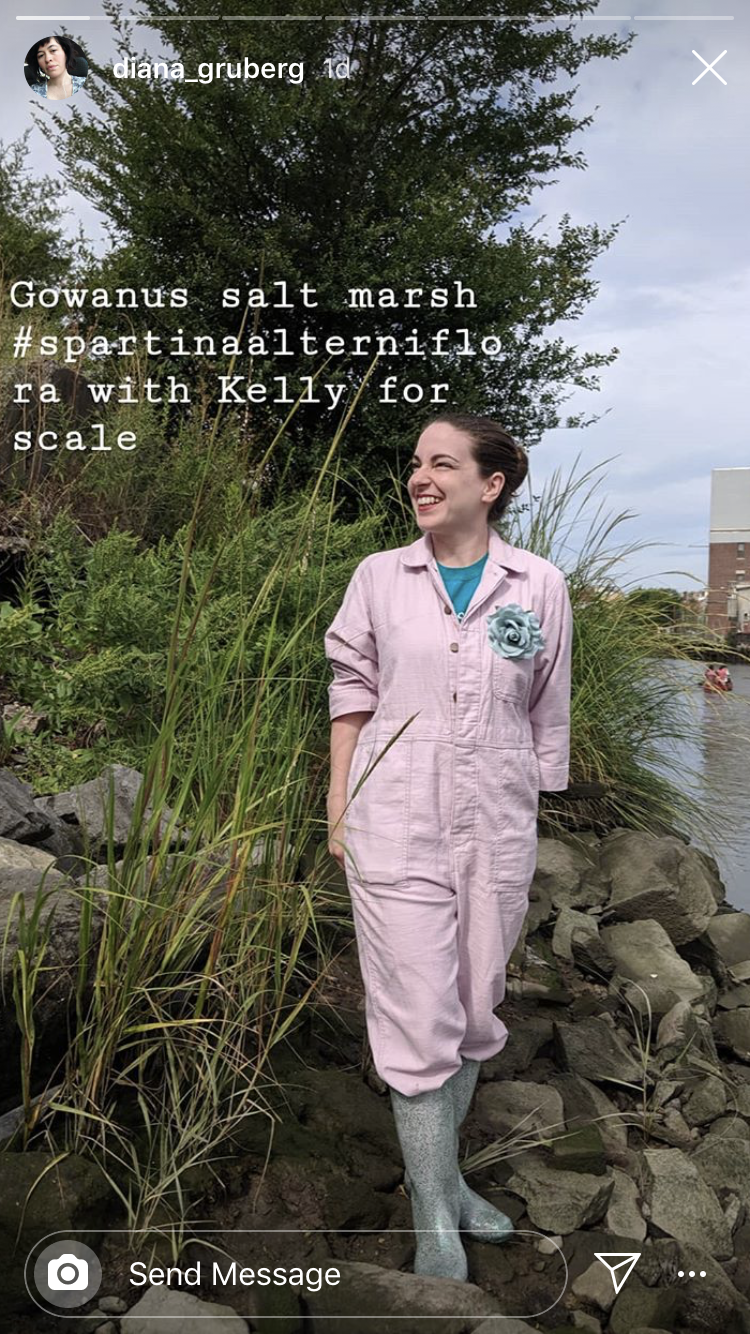 Kelly O'Donnell wears a pink jumpsuit and boots. She is standing in rocks at the Gowanus Salt Marsh.