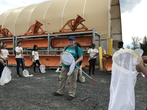 Sarah Kornbluth wears a teal BioBlitz shirt, beige pants, and a blue baseball cap. She is holding an insect net and standing in the Gowanus Canal Salt Lot. Macaulay students holding nets are in a circle around her.