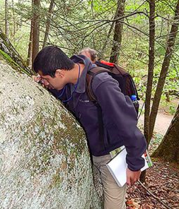 Christian Liriano, wearing a dark blue jacket and a backpack, leans on a rock to examine it closely with a magnifier.