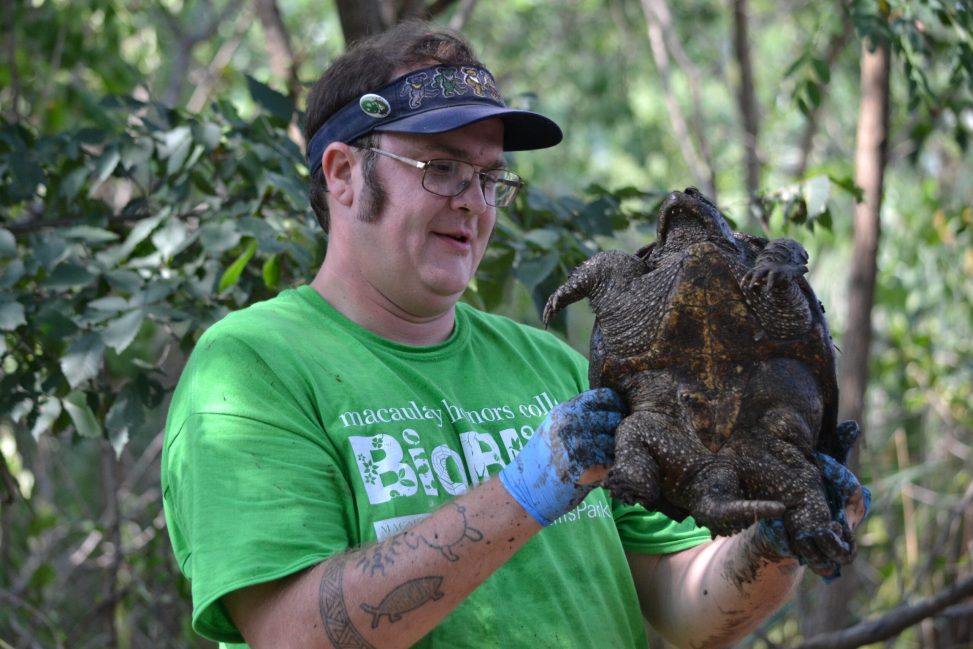 Seth Wollney wears a green BioBlitz Tshirt and a sun visor. He is at Fresh Kills Park and holding a snapping turtle.