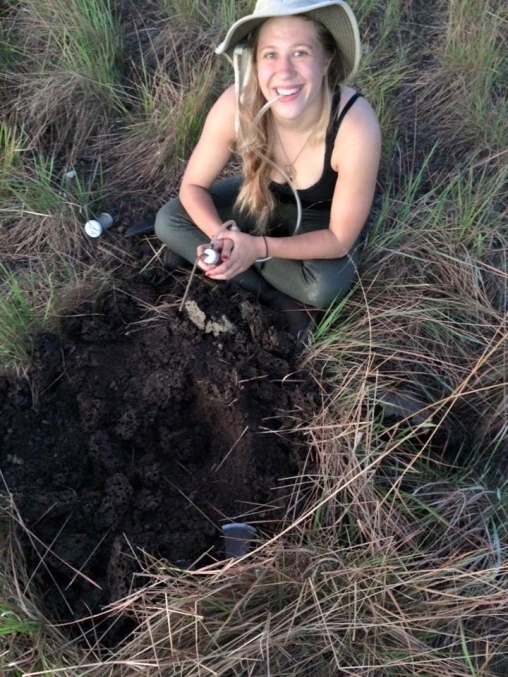 Megan Wilson is a white woman with blonde hair. She is wearing a black shirt and beige hat sits near a termite mound with an aspirator.