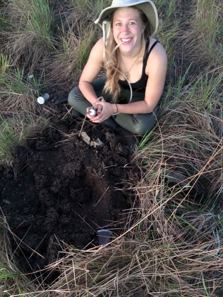 Megan Wilson is a white woman with blonde hair. She is wearing a black shirt and beige hat sits near a termite mound with an aspirator.