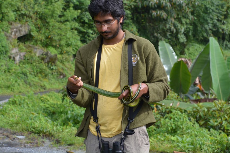 Gautam Surya wears a yellow shirt and khaki jacket. He has binoculars around his neck and is holding a green snake.