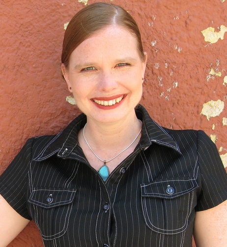 Adrienne Brundage is a white woman with strawberry blonde hair. She is wearing a black shirtdress, red lipstick, and a turquoise necklack.