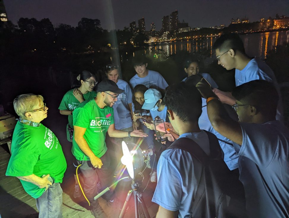 A group of students in blue shirts and scientists in green shirts look at a small insect being held by one of the scientists. They are standing around a bright light used to attract insects.