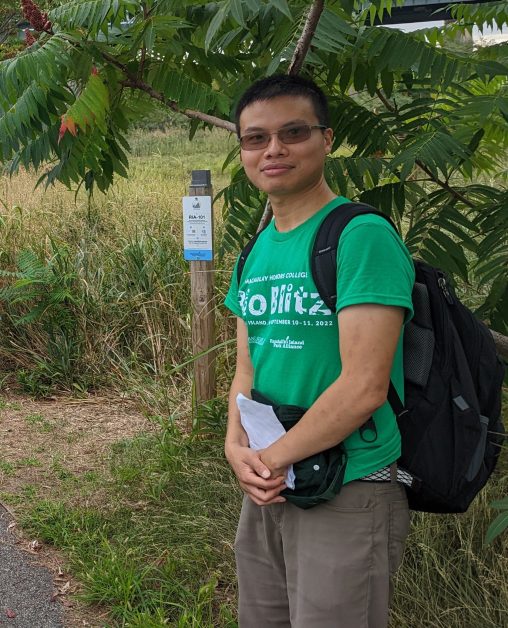 Zihao Wang wearing a green BioBlitz t-shirt and a black backpack standing in front of a trail marker and large sumac shrub.