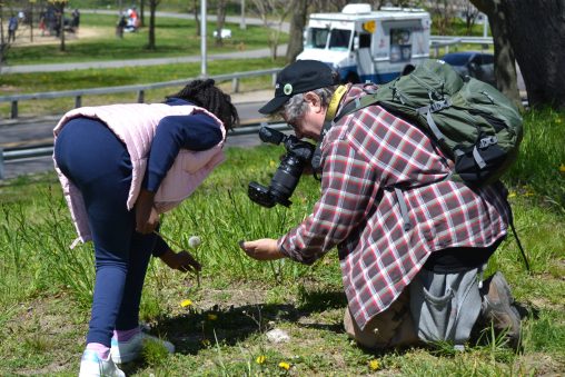 Chris Kreussling wearing a plaid shirt and green backpack kneels down with camera and shows a young girl wearing a pink vest and blue pants a flower.
