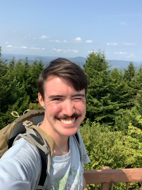 Eli Denzer standing in front of a forest landscape wearing a light blue shirt and backpack.