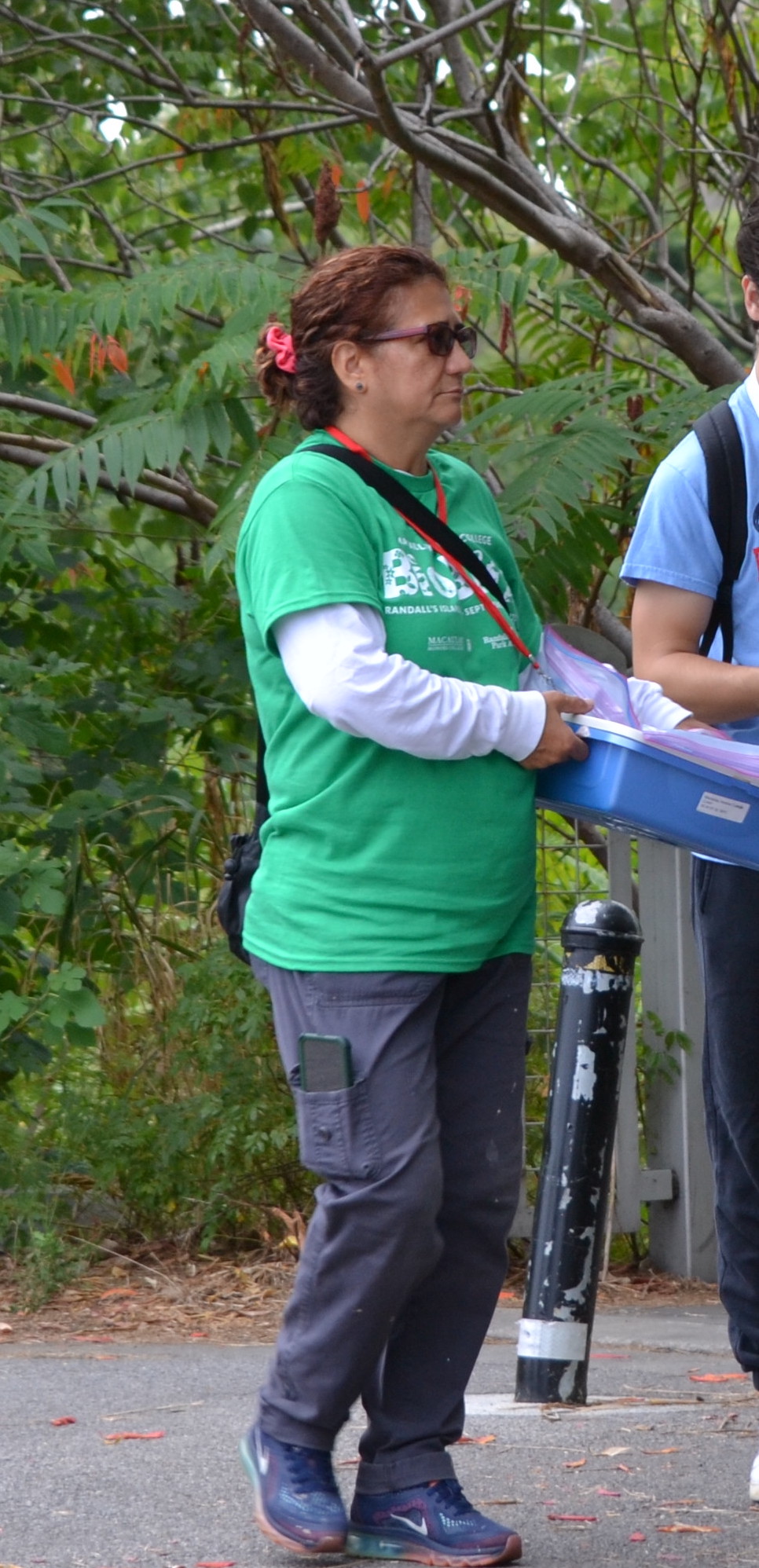Olga Calderon wearing a green BioBlitz shirt and carrying a tray of equipment in front of a sumac.