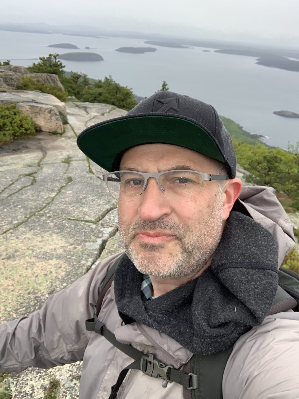 A selfie of Theodore Muth standing on a rocky cliff overlooking a lake. He's wearing glasses, a black baseball cap, and a gray jacket.
