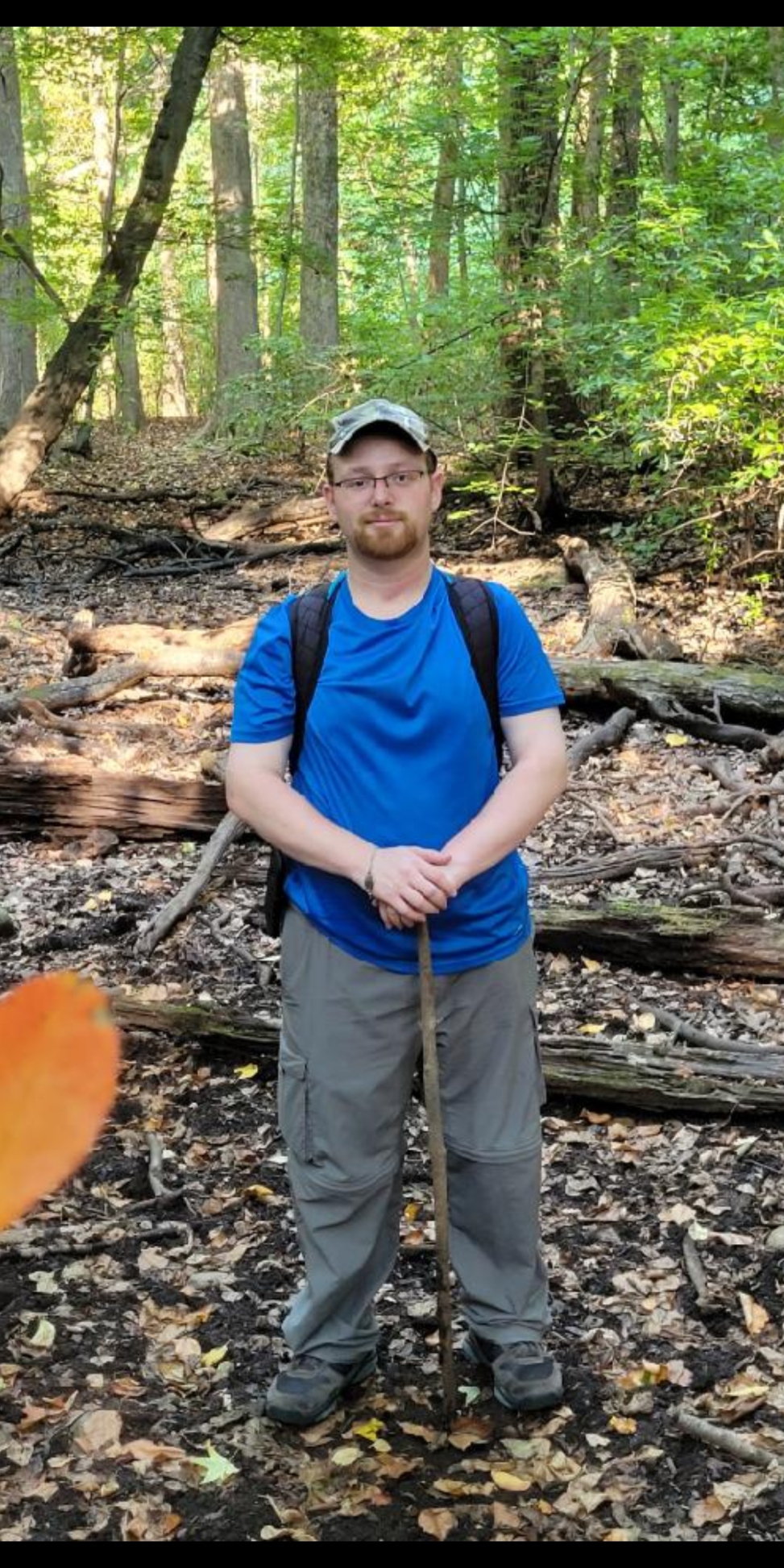 Nickolas Dubin in a baseball cap, blue t-shirt, and gray pants standing at the bottom of a wooded hill.