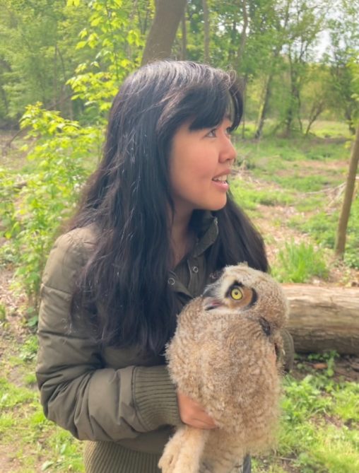 Jacqueline Liang standing in a wooded area wearing a coat. She is holding a very fuzzy owl.
