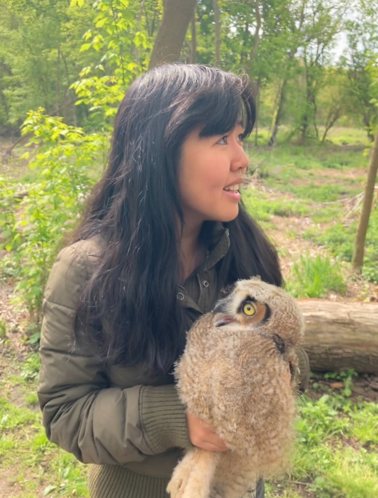 Jacqueline Liang standing in a wooded area wearing a coat. She is holding a very fuzzy owl.
