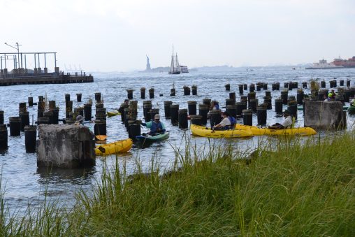 People in yellow kayaks around old shipping piers. Statue of Liberty in the background.
