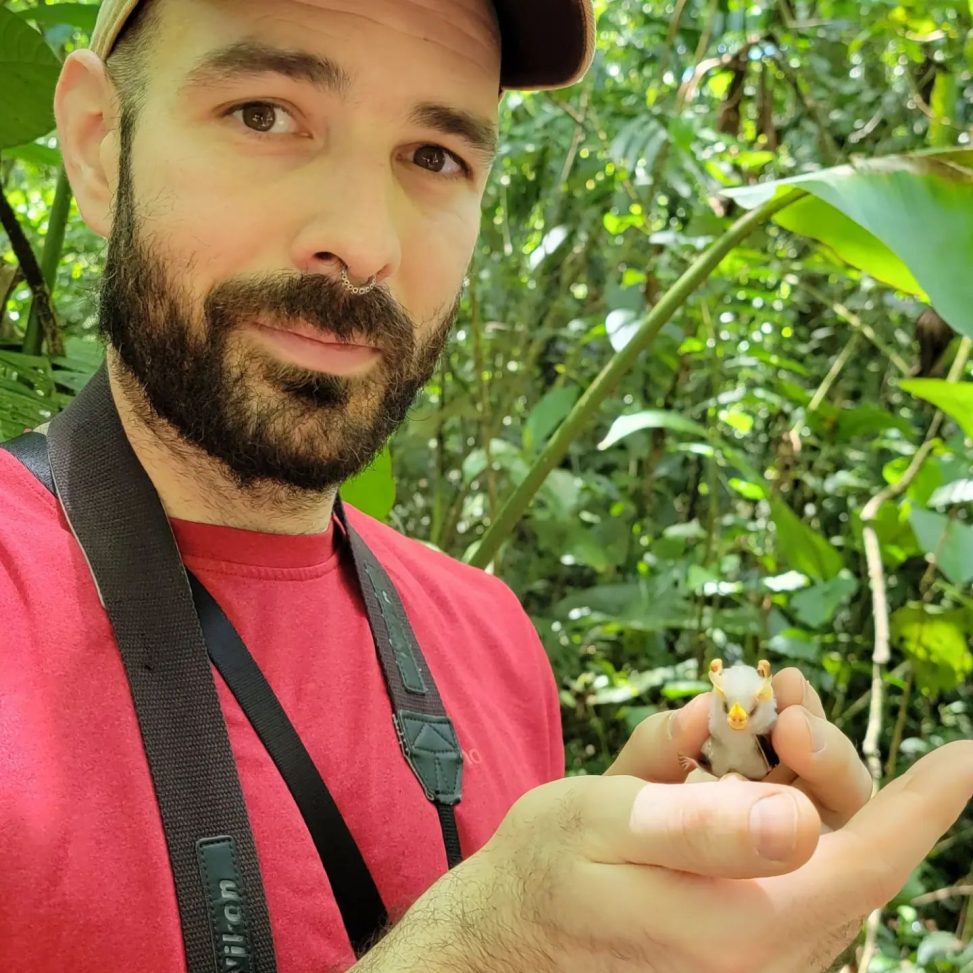 Nic Comparato standing in front of a leafy background and holding a bat that is about the size of one of their fingers.