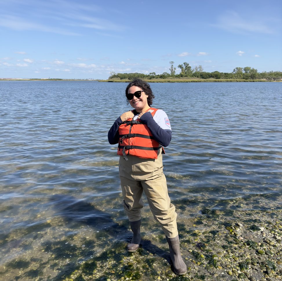 Isabella Torres wearing hip waders and a life vest standing on a rocky shore.