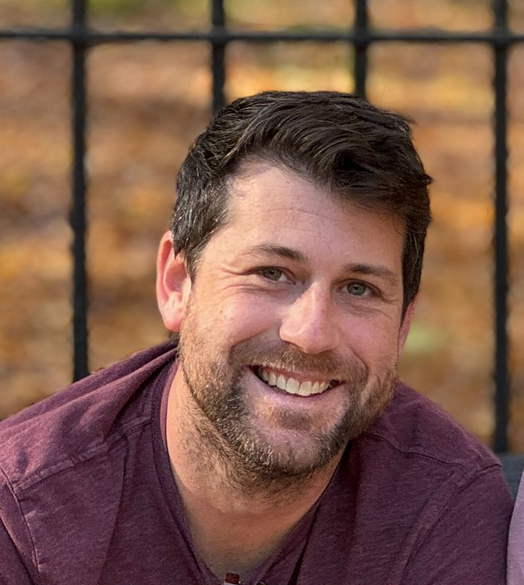 This is a headshot of Brandon Ely. He's wearing a burgundy shirt and is in front of a metal fence.