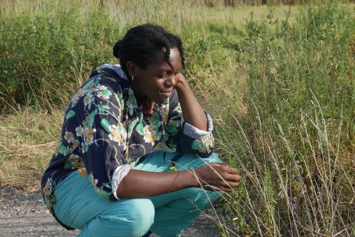 Maren Fossi on a path through a grassy wetland habitat. She is wearing green pants, a black and green printed shirt and is crouching down to look at a plant.
