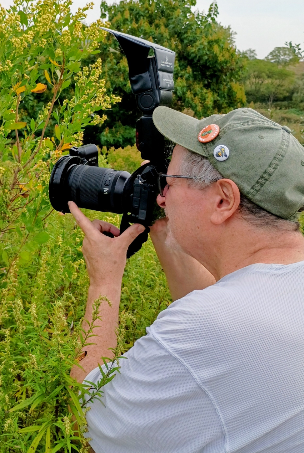 Photo of Chris Kreussling taken over the shoulder. They are wearing a green hat and light colored shirt and holding up a large black DSLR camera to photograph an insect on a plant.