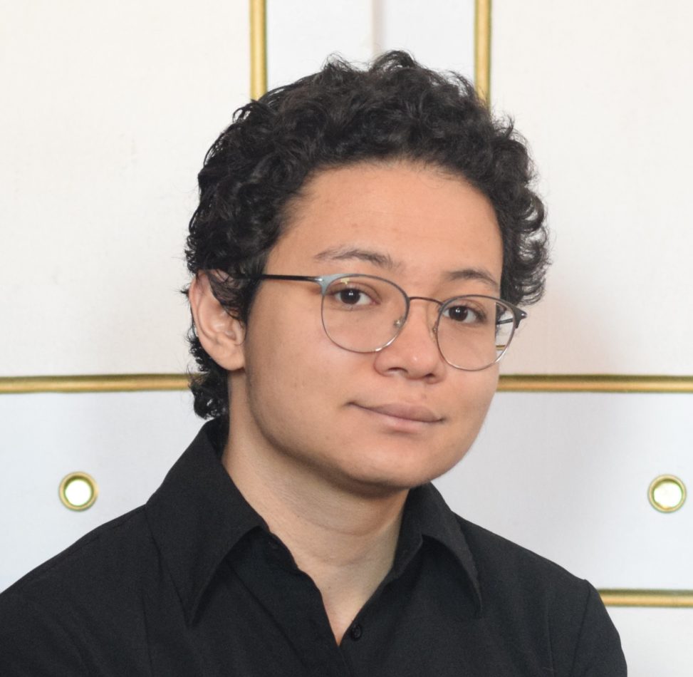 Masami in front of a white wall looking at the camera with a smile. He's wearing glasses and a dark colored shirt.