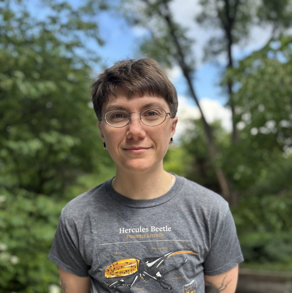 Photo of Rin standing in the woods. He is wearing glasses, a gray t-shirt with a Hercules beetle on it and looking directly at the camera.