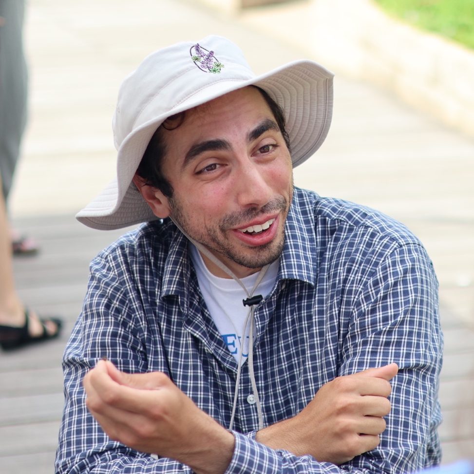 Nick at Lurie Garden on a wooded path. He's wearing a floppy light colored hat and a blue plaid shirt.