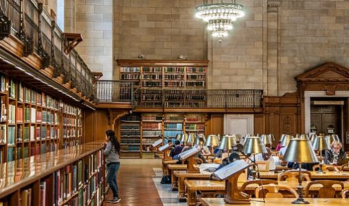 Reading Room at the main branch of the New York Public Library. Long tables with lamps and a person browsing books around the room's perimeter.