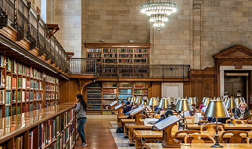 Reading Room at the main branch of the New York Public Library. Long tables with lamps and a person browsing books around the room's perimeter.