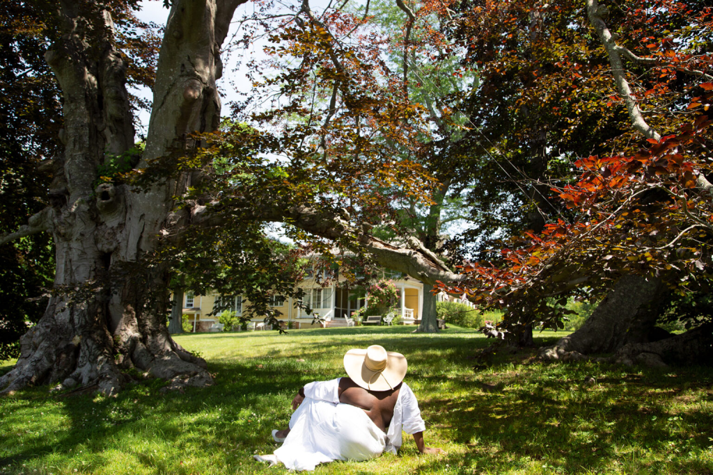 Woman in a white dress and sunhat sits in the grass looking at house
