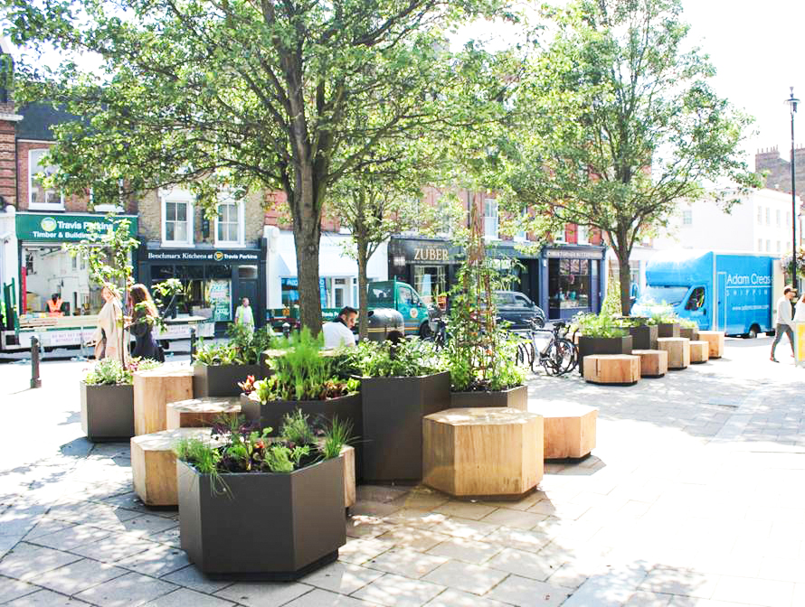 A streetscape features geometric planters and wooden seating surrounded by trees, with shops and a blue delivery truck in the background. People are walking and standing in the urban setting.