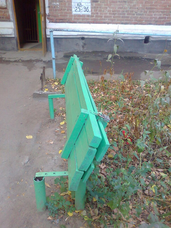 A green wooden bench is folded upright and padlocked in an outdoor setting with autumn leaves on the ground. The bench appears to be in front of a brick building with an open doorway.