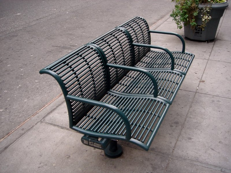 A dark green, slatted metal park bench with three seats sits on a concrete sidewalk next to a street. A planter with greenery is visible in the background.