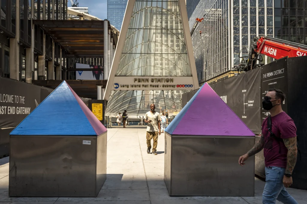 Two pyramid sculptures with blue and pink tops stand in front of the Penn Station entrance. A man wearing a mask walks past the sculptures.