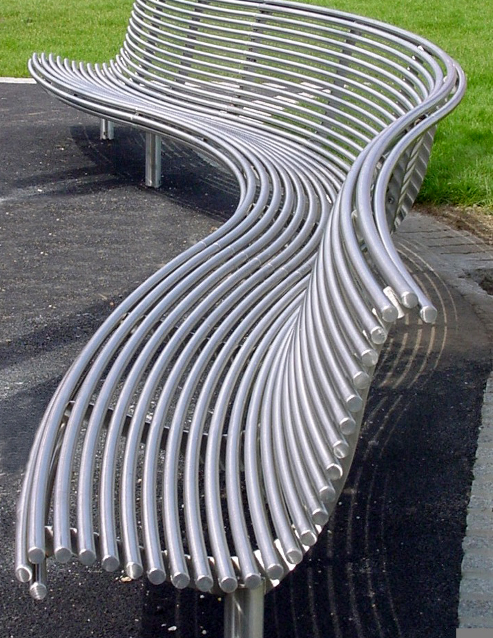 A modern, curved metal bench with many parallel, wavy slats is shown in a park. The bench is made of shiny silver metal and is supported by a few legs.