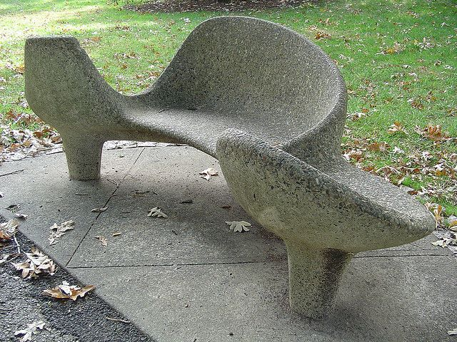A grey concrete bench with a sculptural, flowing design sits on a paved surface. The bench is surrounded by grass and fallen leaves.